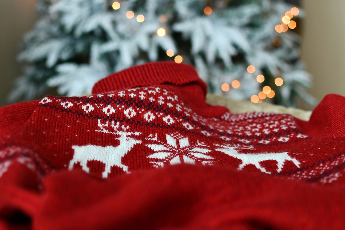 Röd stickad tröja med vita renar och snöflingor i mönsterband, framför en julgran med ljus
